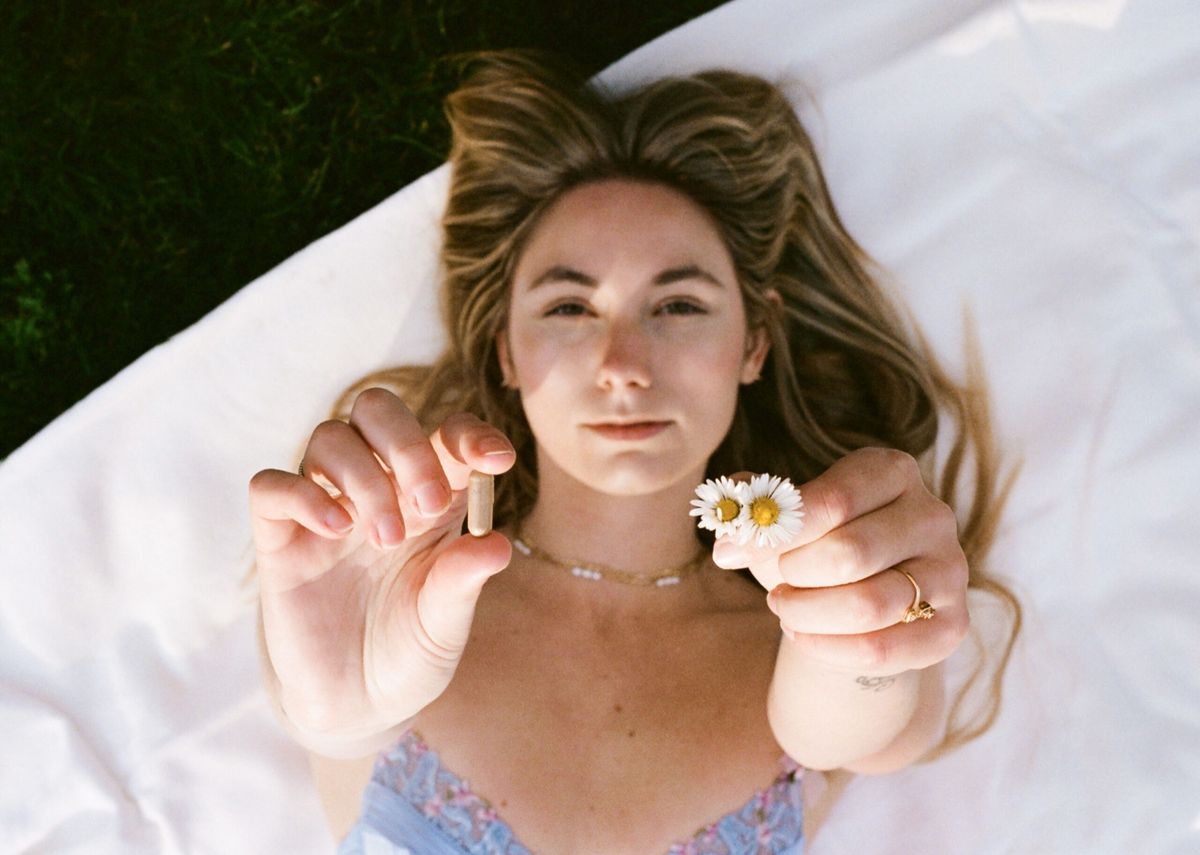 A woman lying on white linen holding up a capsule and a daisy.