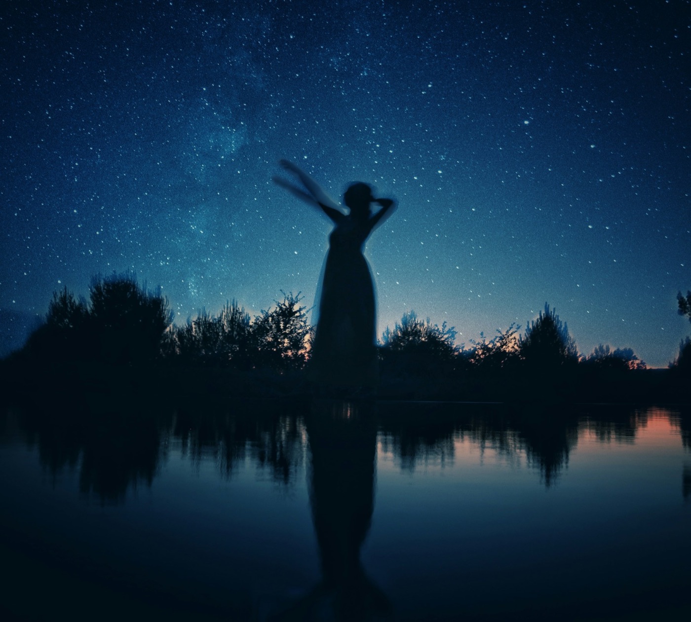 A woman standing on a rock beneath a starry sky reflected in a still lake